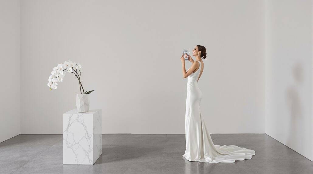 Bride in a white wedding gown taking photos with her phone in a minimalist setting.