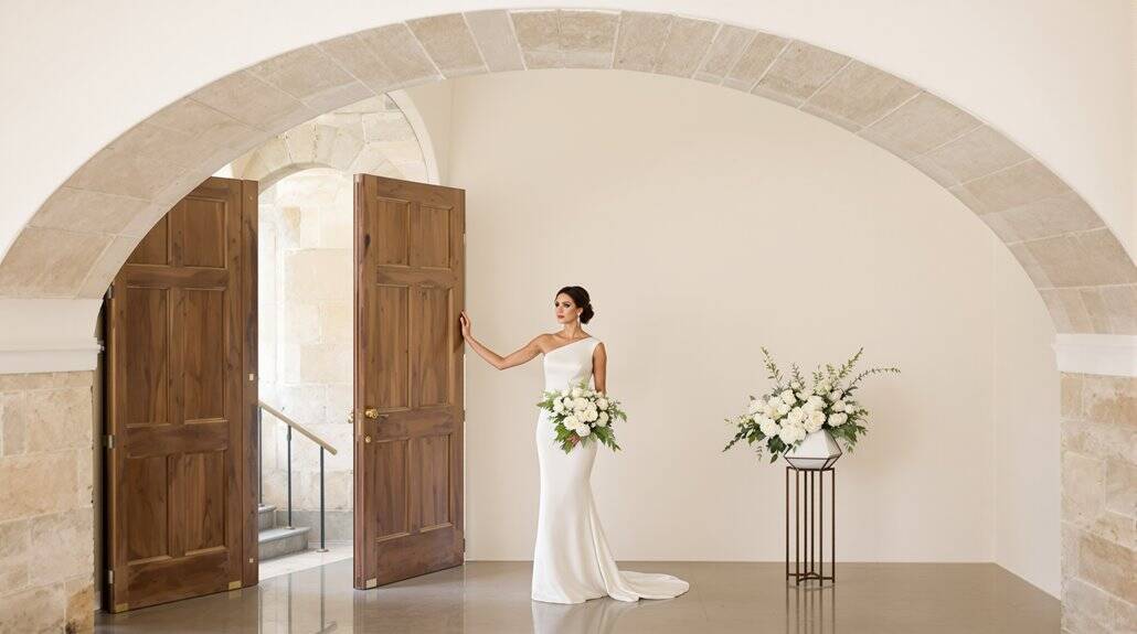 Bride in elegant wedding dress holding bouquet near open wooden doors.